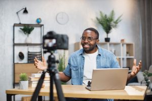 Young freelancer using a modern laptop and digital camera for a split-face video recording at home.